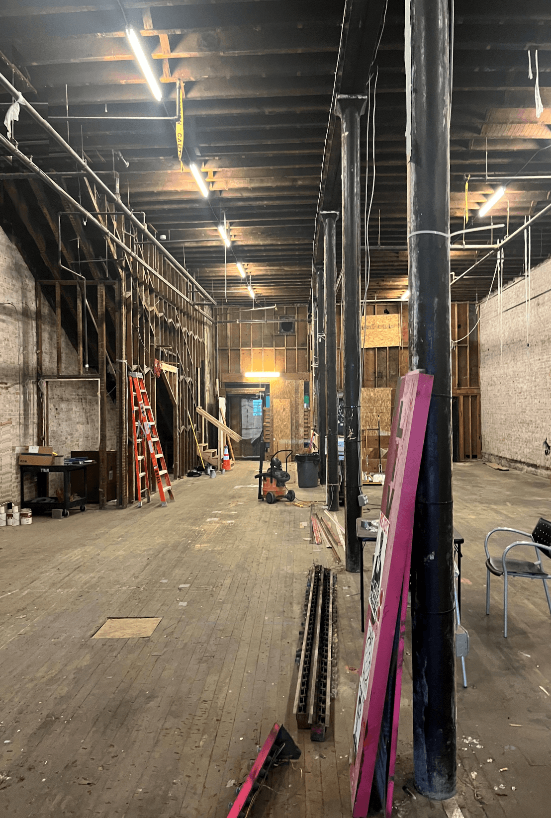 Standing at the back of the gutted Lusty Lady, looking toward the front: original wood ceiling beams, exposed brick on both sides, construction lights overhead, a ladder, a recycling can, and a folded chair in the dust