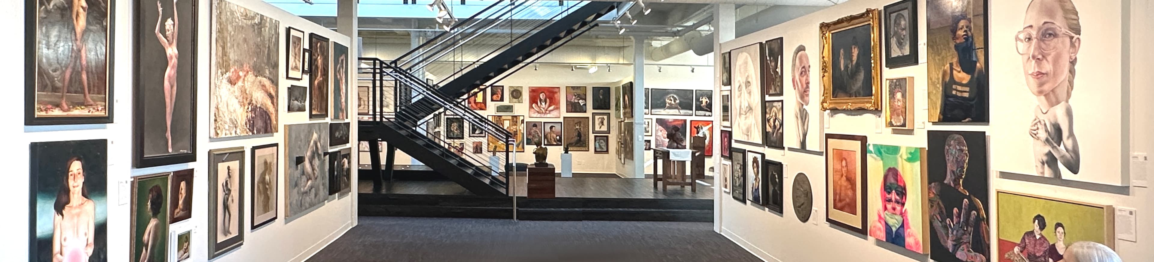 The Art Love Salon downstairs gallery: salon-style hang of contemporary paintings on white walls in a long, light-filled space, with an open staircase up to the mezzanine and a wide central aisle