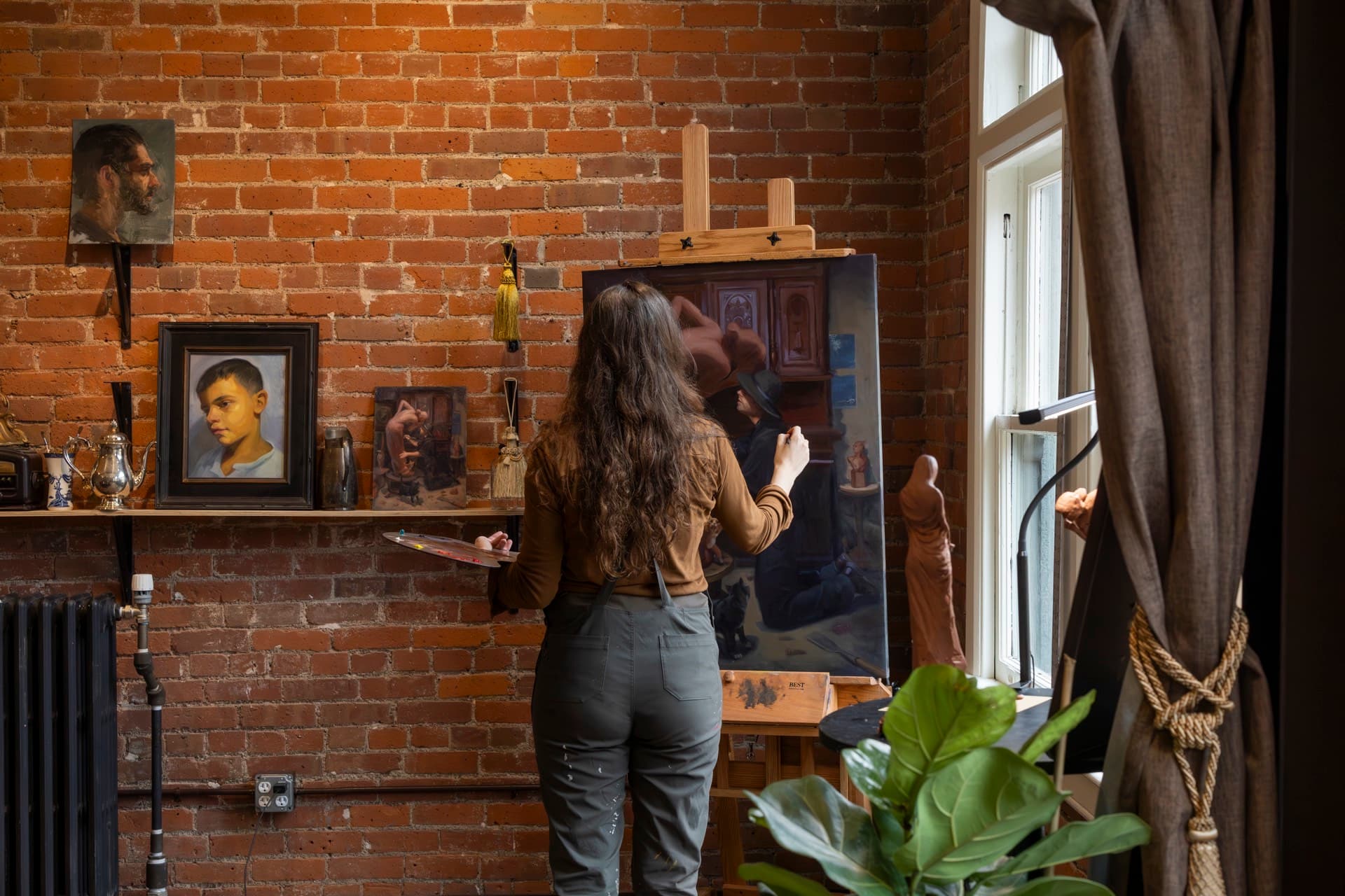 A painter working at the easel from behind, with framed studies and a small portrait on a shelf against the brick wall of her Occidental studio.